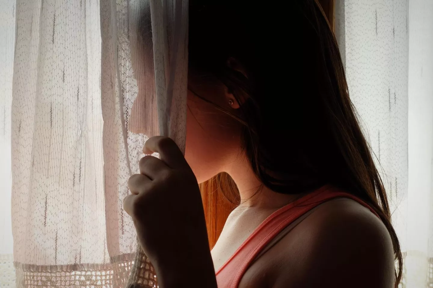 Adolescente mujer de perfil, mirando por una ventana. No se ve su cara porque sostiene una cortina que la tapa.