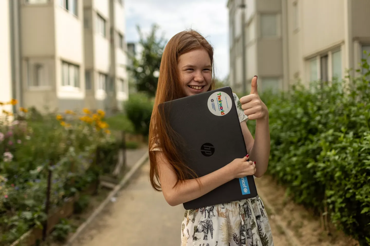 Adolescente sosteniendo una laptop en un brazo, y con la otra mano hace una se&ntilde;a de pulgar para arriba. Est&aacute; sonriendo.