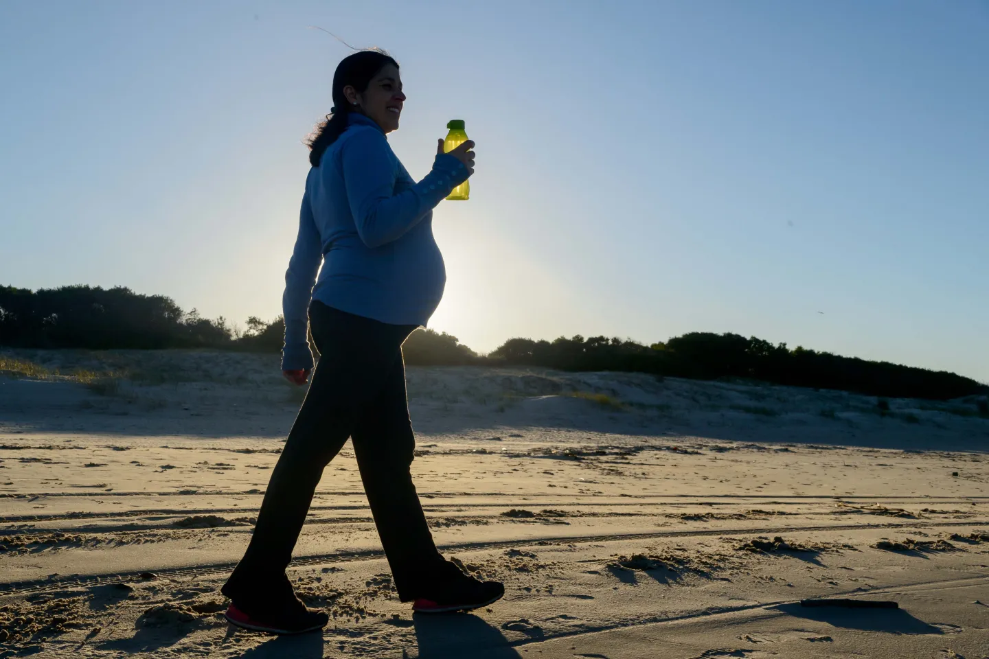 Mujer embarazada caminando en la playa con botella de agua