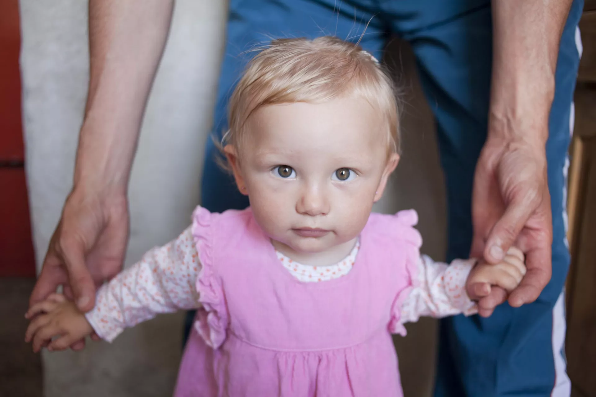 Ni&ntilde;a de aproximadamente un a&ntilde;o, parada con su padre por detr&aacute;s, que la toma de las manos.