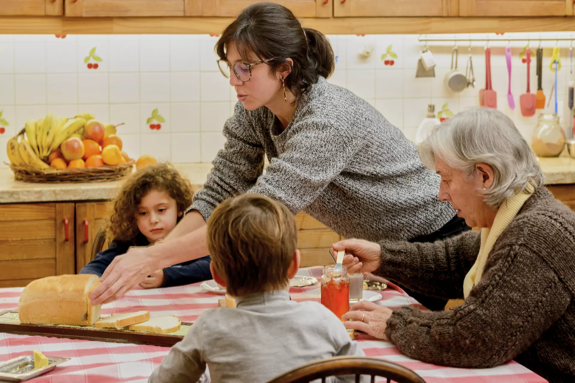 Mujer cortando pan sobre mesa, donde est&aacute;n sentados un ni&ntilde;o, una ni&ntilde;a y una se&ntilde;ora mayor.