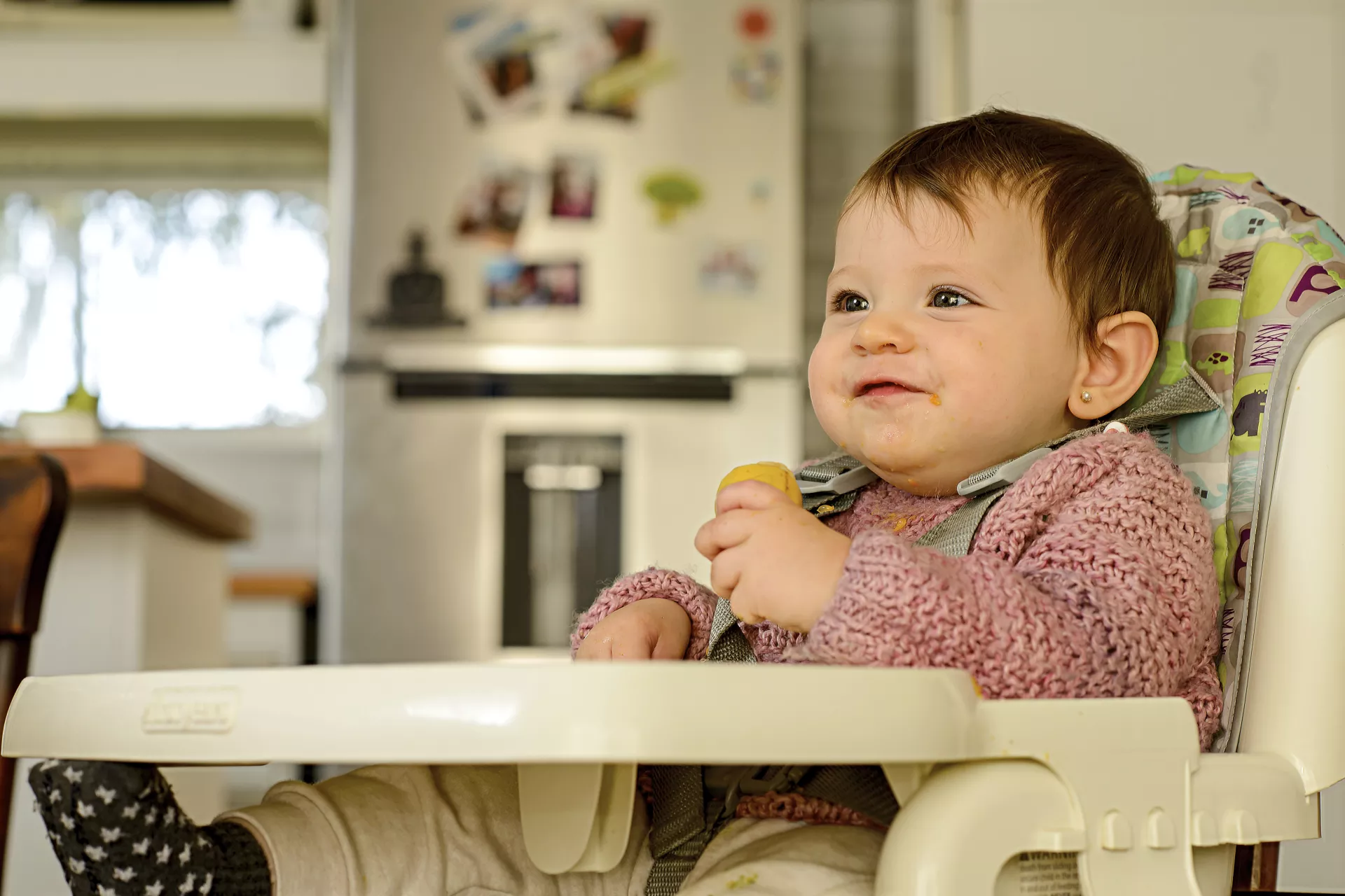 Beb&eacute; de unos 8 meses, sentada en sillita de comer, agarrando una fruta con la mano.