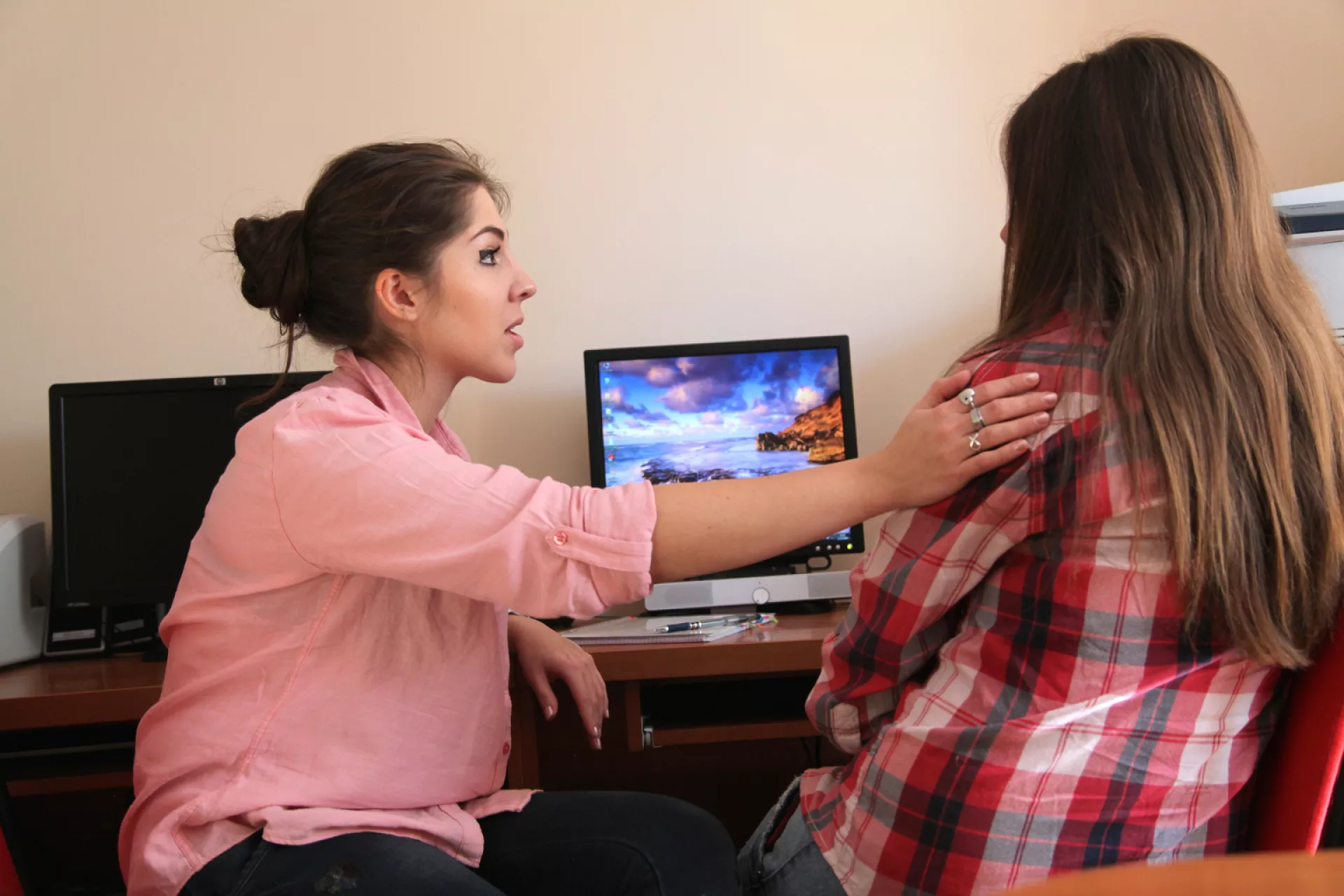 Adulta poni&eacute;ndole la mano en el hombro a adolescente mujer que est&aacute; dada vuelta. Adelante de la adolescente hay una computadora.