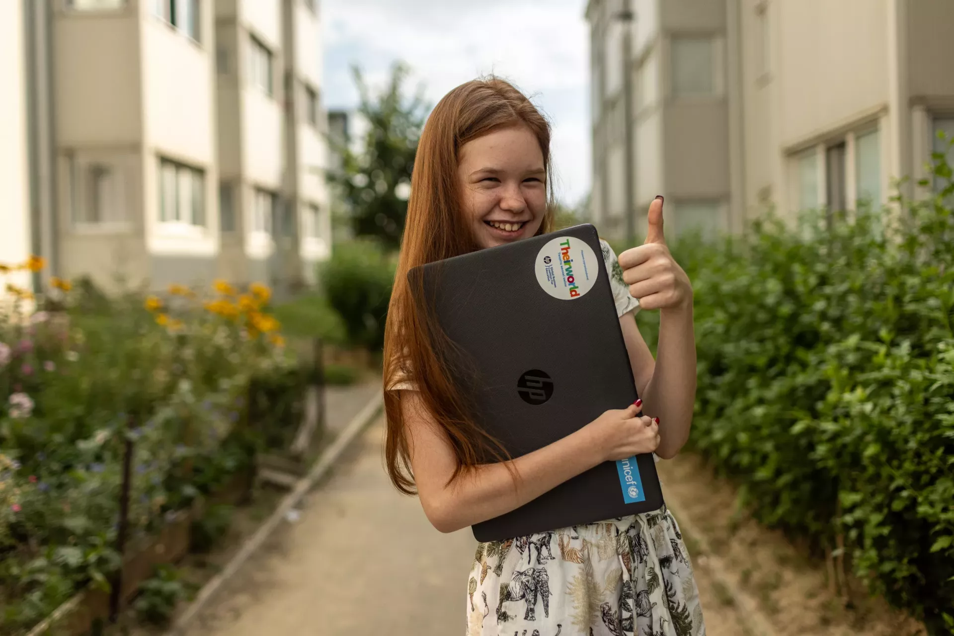 Adolescente sosteniendo una laptop en un brazo, y con la otra mano hace una se&ntilde;a de pulgar para arriba. Est&aacute; sonriendo.