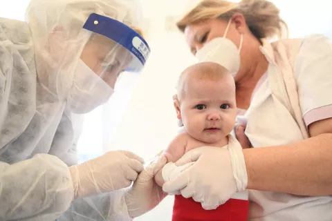 Bebé siendo vacunado por una persona con vicera transparente y tapabocas. La mujer que sostiene al bebé también tiene tapabocas y guantes.