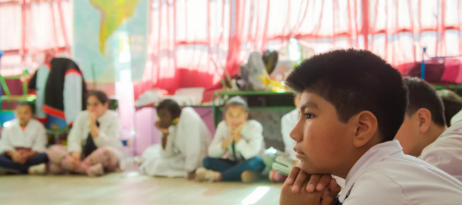 Ni&ntilde;o en una ronda, sentado en el piso junto con otros ni&ntilde;os. Todos est&aacute;n de t&uacute;nica en una escuela.