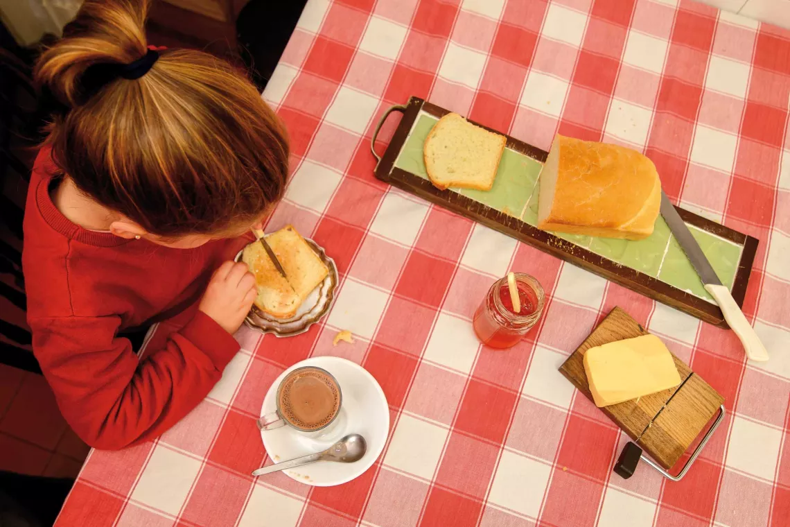 Ni&ntilde;a, vista desde arriba, untando un pan casero. En la mesa est&aacute; el resto del pan, mermelada, manteca, y una leche chocolatada.