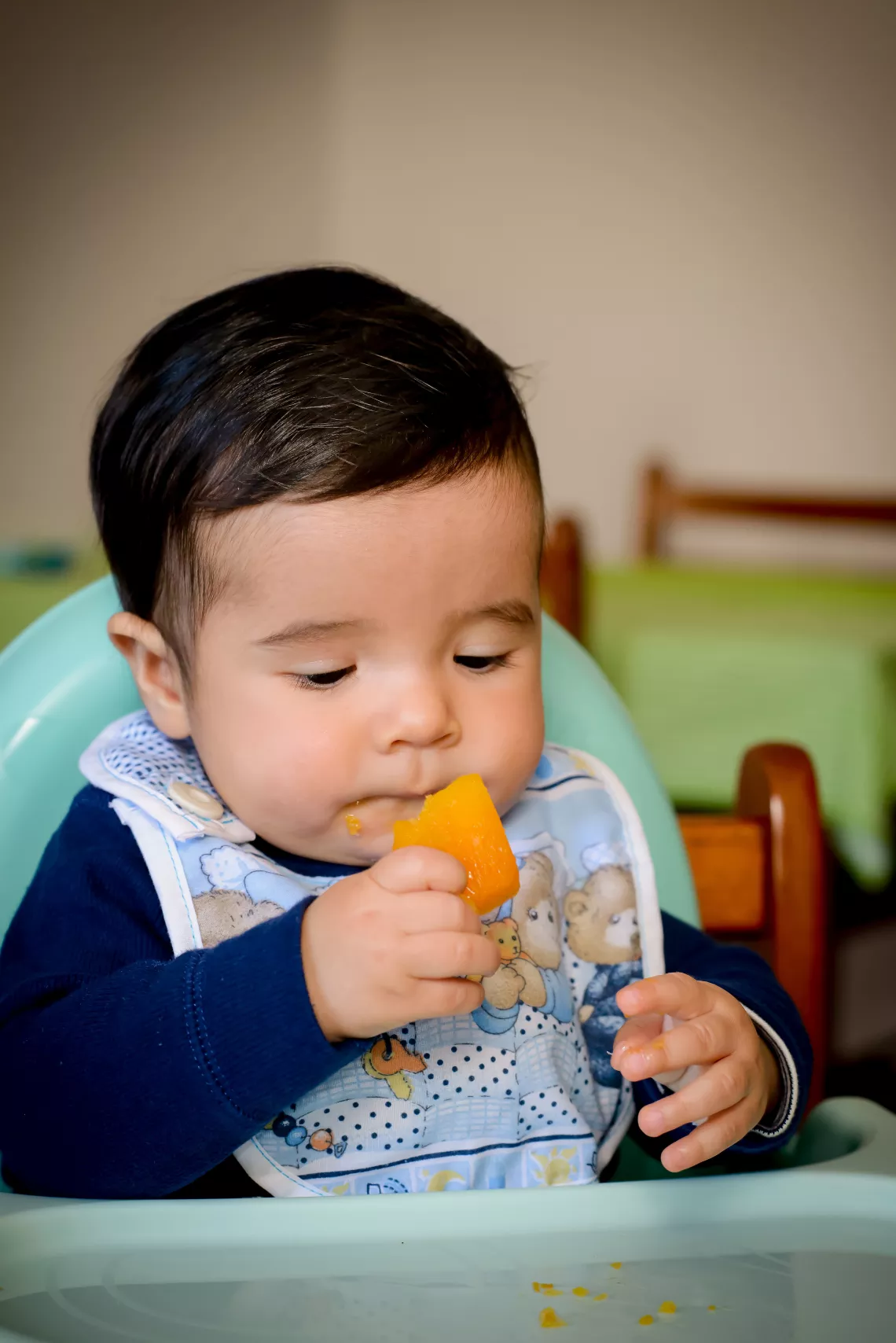 Ni&ntilde;o peque&ntilde;o, sentado en sillita de comer, tomando un trozo de calabac&iacute;n con una mano.