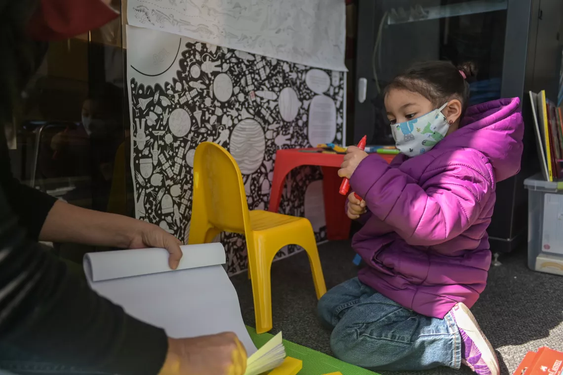 Ni&ntilde;a de unos 3 a&ntilde;os, con tapabocas, jugando con crayolas, en espacio acondicionado para juego de ni&ntilde;os.