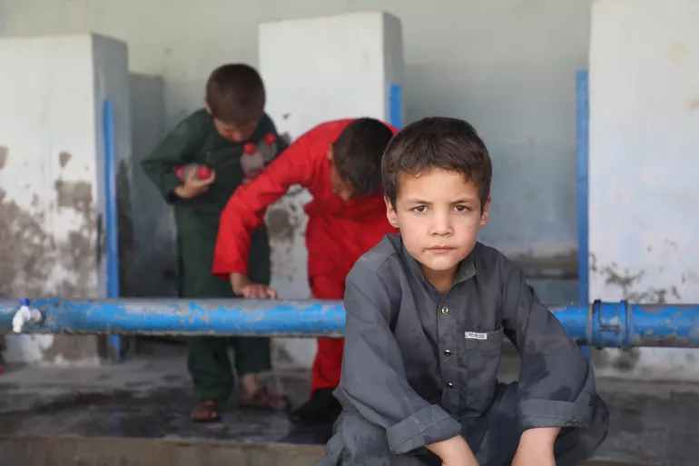 Ni&ntilde;o en cuclillas mirando a c&aacute;mara. Detr&aacute;s de &eacute;l hay otros dos ni&ntilde;os inspeccionando un ca&ntilde;o y con algunas botellas en sus manos.