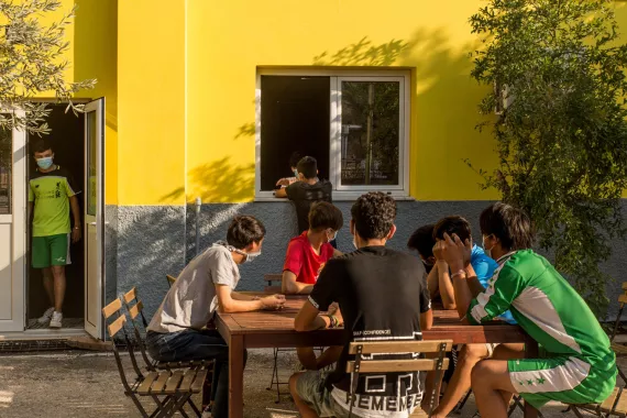 Grupo de adolescentes varones, con tapabocas, sentados al aire libre alrededor de una mesa