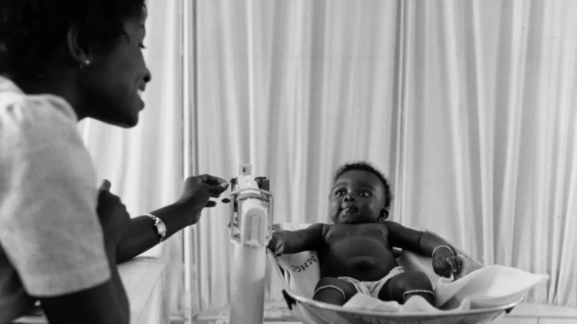 En 1960 en Ghana, una partera pesa a un ni&ntilde;o en una balanza como parte de un plan integral de salud rural a largo plazo, promovido por UNICEF, que enfatizaba en la capacitaci&oacute;n del personal m&eacute;dico en Tuma.