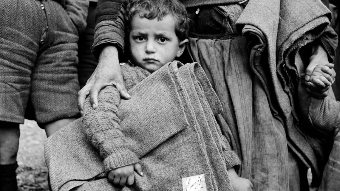 Alrededor de 1950, en la ciudad noroccidental de Castoria, Grecia, un ni&ntilde;o sostiene una manta proporcionada por UNICEF, agarrando la falda de una mujer.
