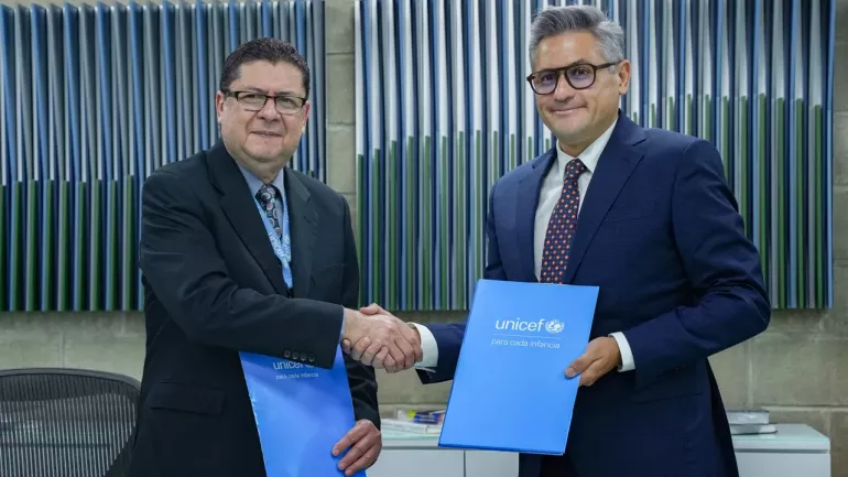 El representante adjunto de UNICEF Venezuela, Jos&eacute; Ram&oacute;n Espinoza y el director ejecutivo de El Sistema posan ante la c&aacute;mara d&aacute;ndose la mano y mostrando unas carpetas azules con el logo de UNICEF