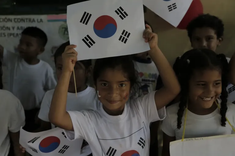 Samantha (6) mira a la c&aacute;mara y levanta una bandera junto a Valerie (6) mientras celebran con sus compa&ntilde;eros de clase la visita a su escuela de Hansang Lee, Encargado de Negocios de la Rep&uacute;blica de Corea en Venezuela, acompa&ntilde;ado por representantes de UNICEF en Maracaibo, estado Zulia, Venezuela, el 11 de junio de 2026.