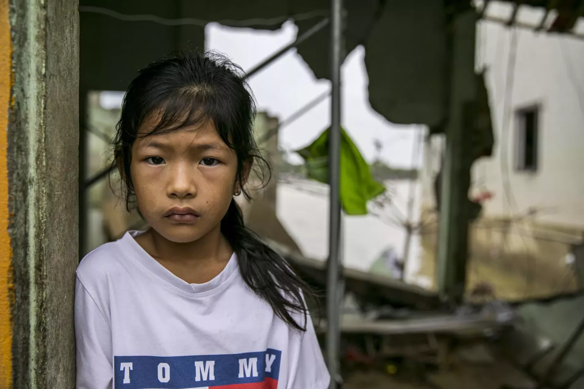 A girl looking straight to us, her eyes filled with despair. Behind her is a destroyed house.