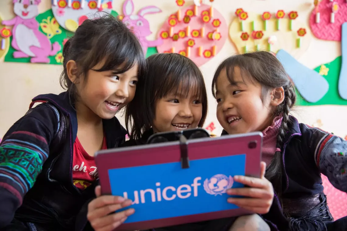 Three little girls smiling, the girl in the middle holds a tablet with UNICEF logo on the back