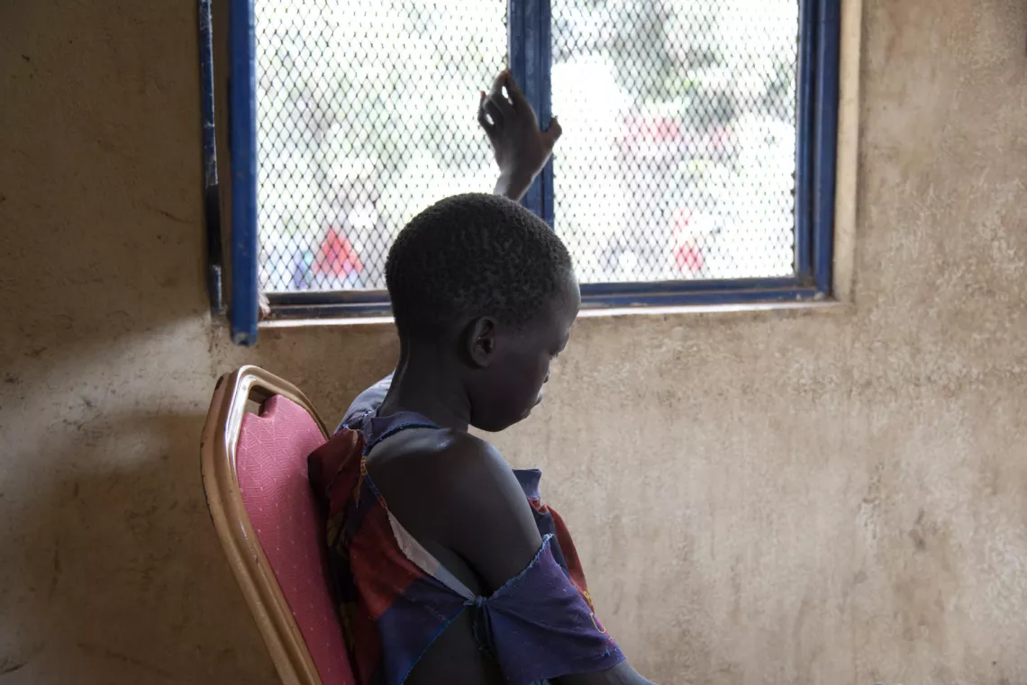 Martin [NAME CHANGED], 13, touches a window in the special protection room of a holding centre for women and children in South Sudan. They are waiting for their cases to come up for court. His Barcelona football t-shirt has huge tears and his shorts are tied around with a piece of string.