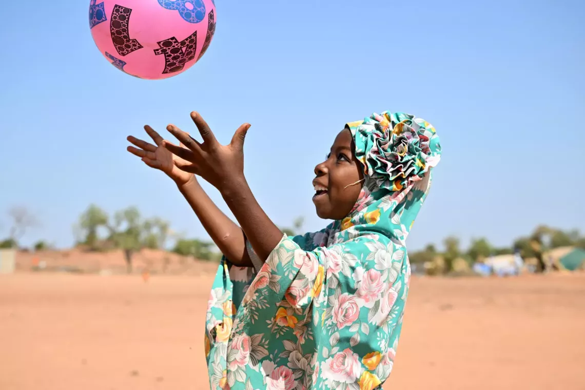 A girl in a floral scarf smiles as she tosses a pink ball into the air. 