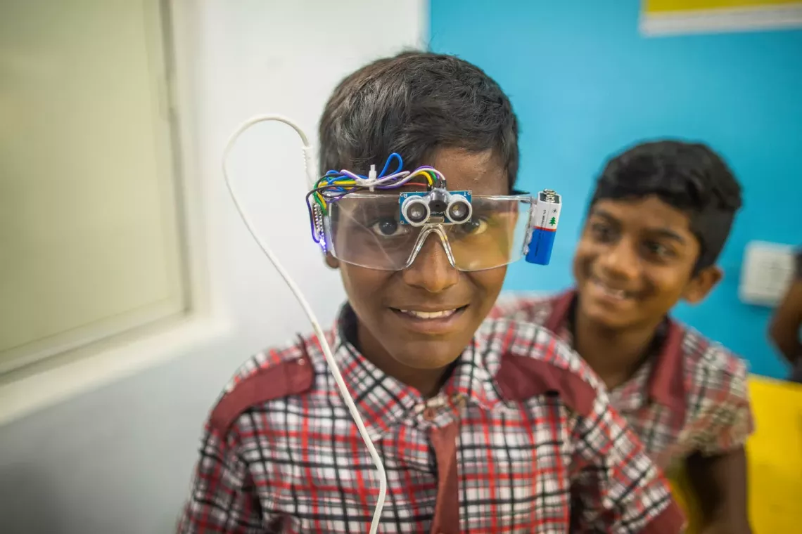 A student wears goggles retrofitted with a battery pack and camera. A peer smiles in the background.