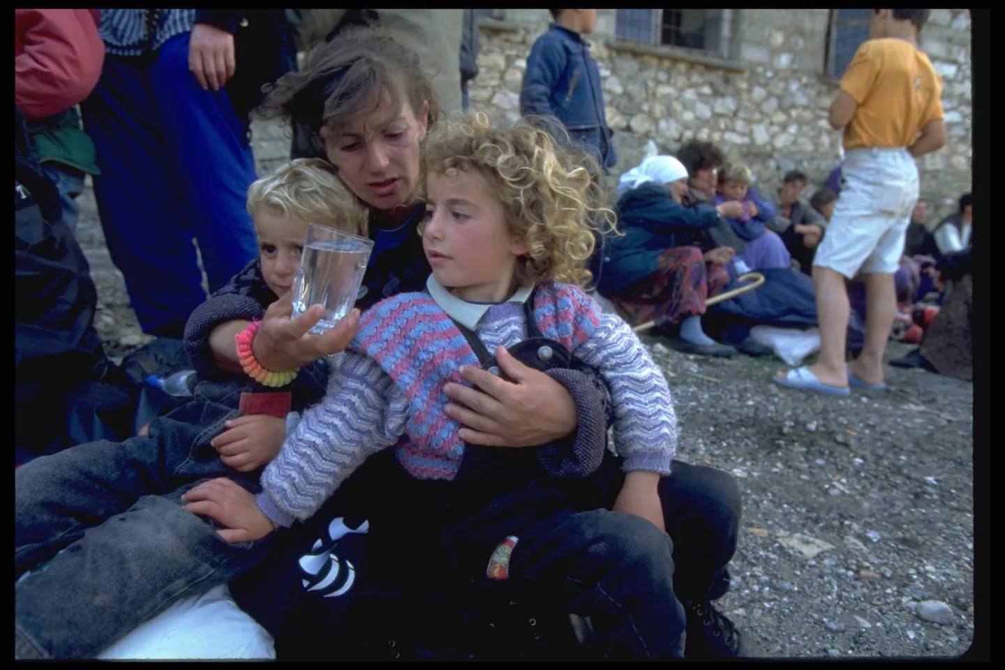 Her two children on her lap, an ethnic Albanian woman from Kosovo offers a glass of water to her daughter as they sit on the ground with other refugees at a commune in the town of Fierze, waiting to be taken by truck to nearby apartment buildings where they will be housed with local families.
