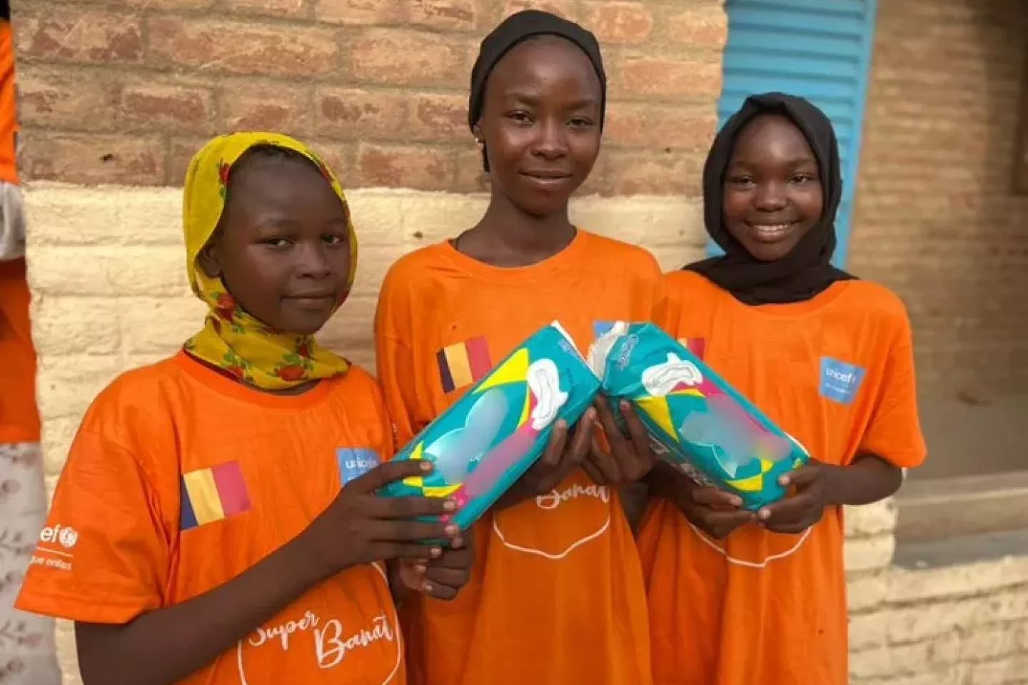Three girls wearing orange t-shirts holding packs of menstrual pads