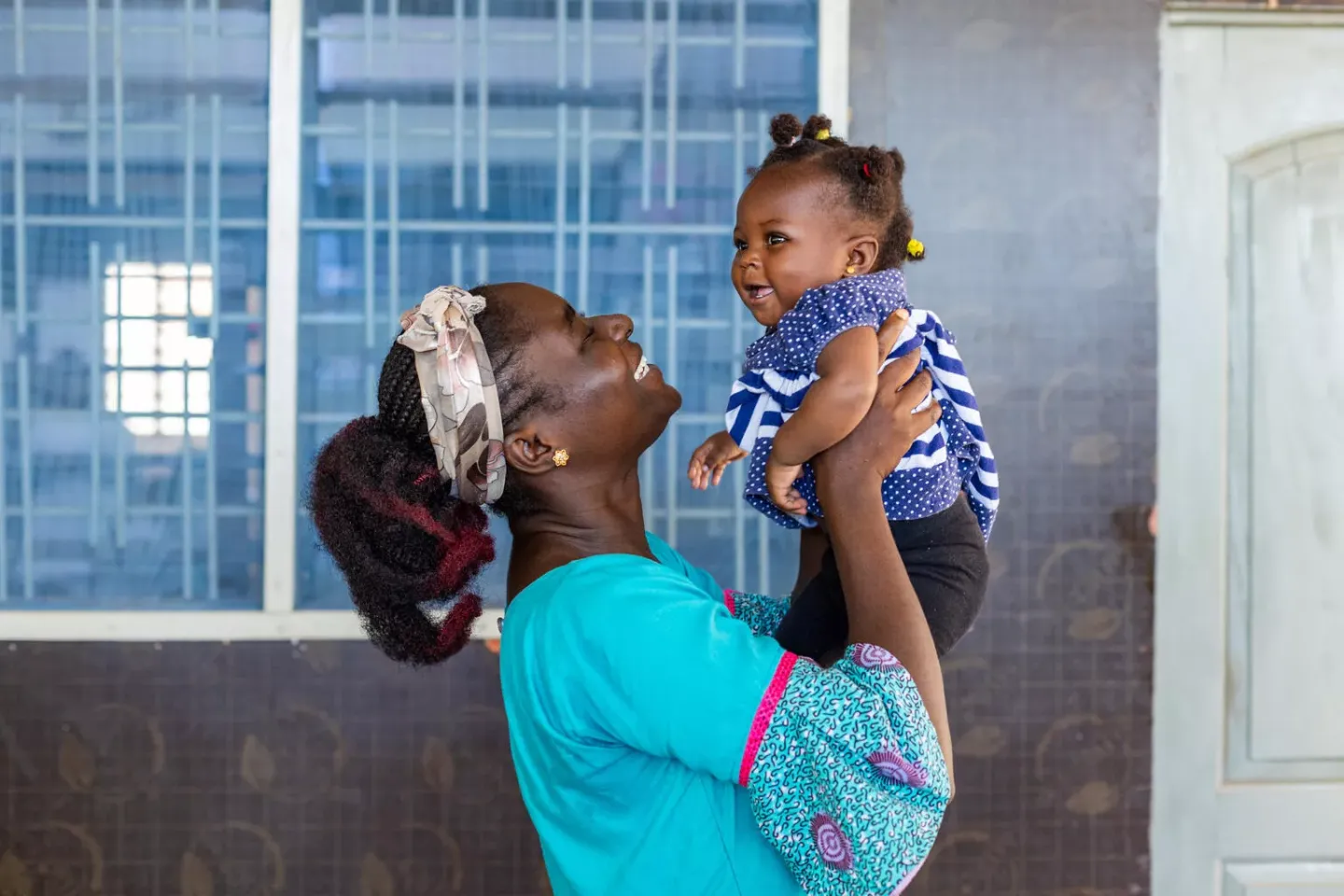 A mother wearing a blue dress smiling as she playfully holds her smiling baby daughter up 