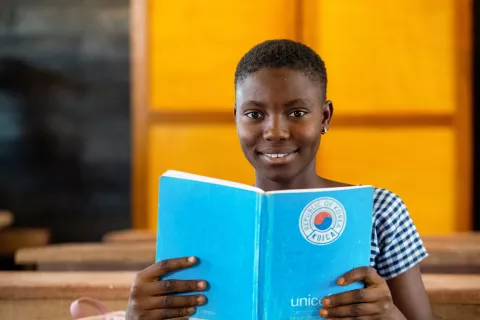 An adolescent girl smiles and holds her textbook in her classroom