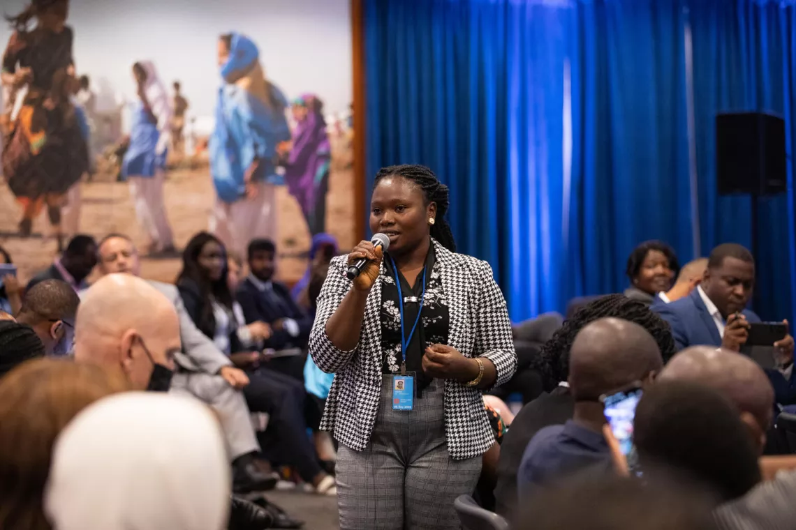 A young woman standing and speaking into a mic in the middle of a seated crowd