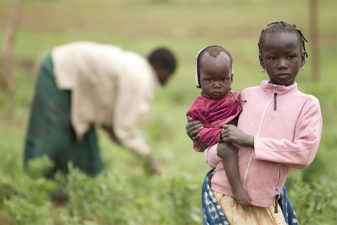 As a woman works behind them, a girl holds her younger sister, standing in a vegetable garden in the village of Noussou, Nord Region.