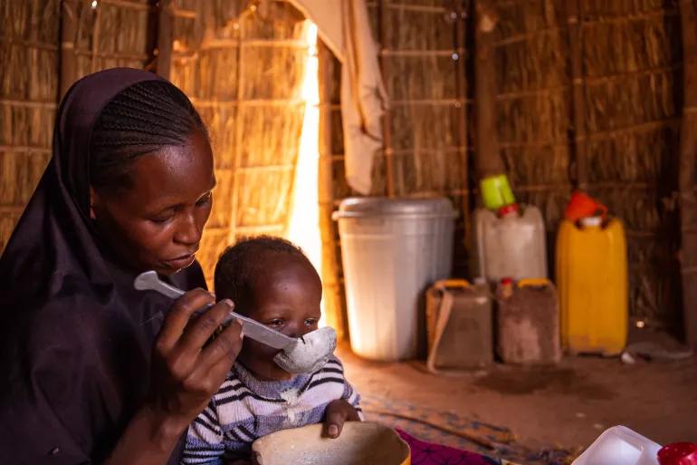 A mother sitting on the floor feeding her child with a spoon