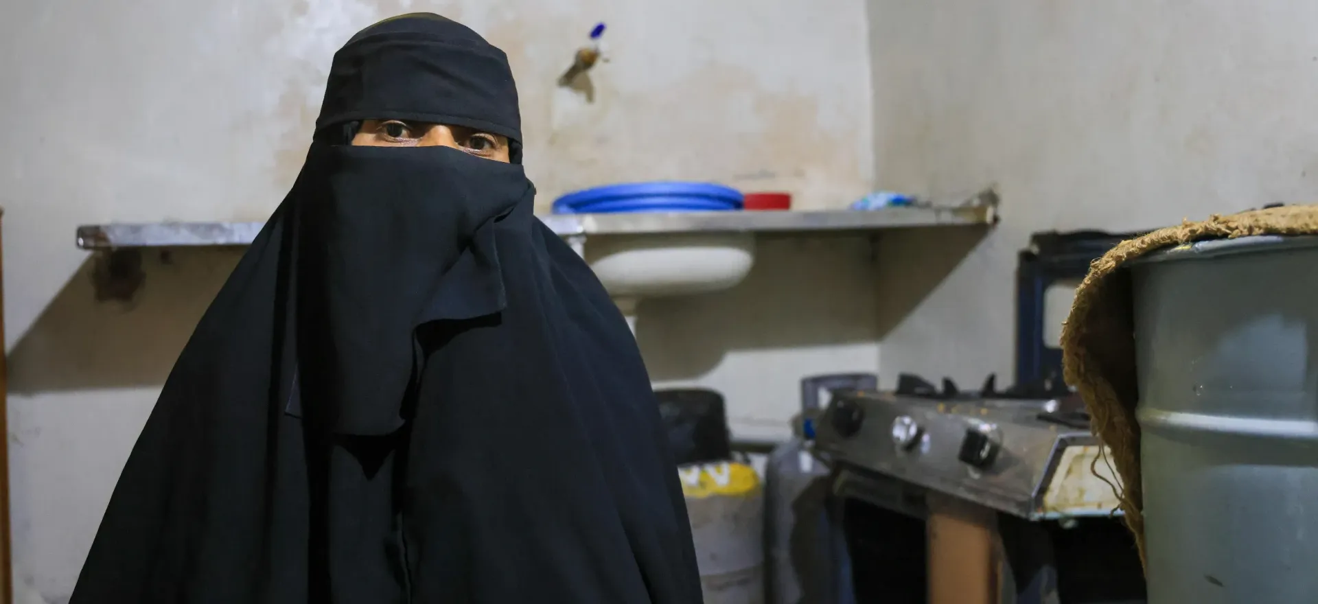 A woman wearing a black niqab standing inside a home kitchen near a sink and gas stove setup.