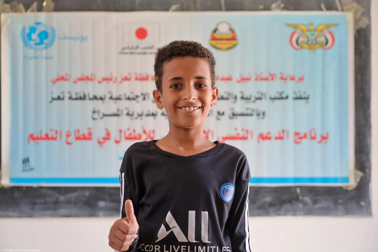 A close-up portrait of the boy giving a thumbs-up gesture while smiling towards the camera.