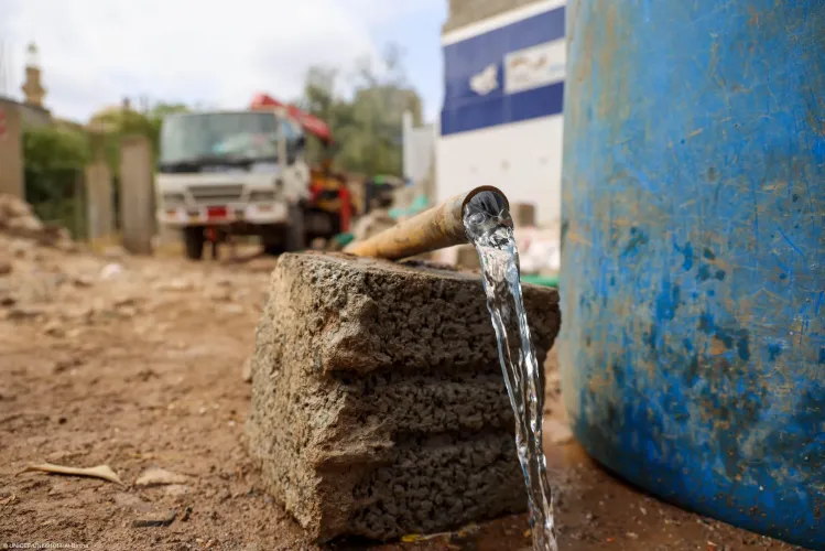 Clean water flowing continuously from an open metal pipe supported by concrete blocks next to a large blue water container