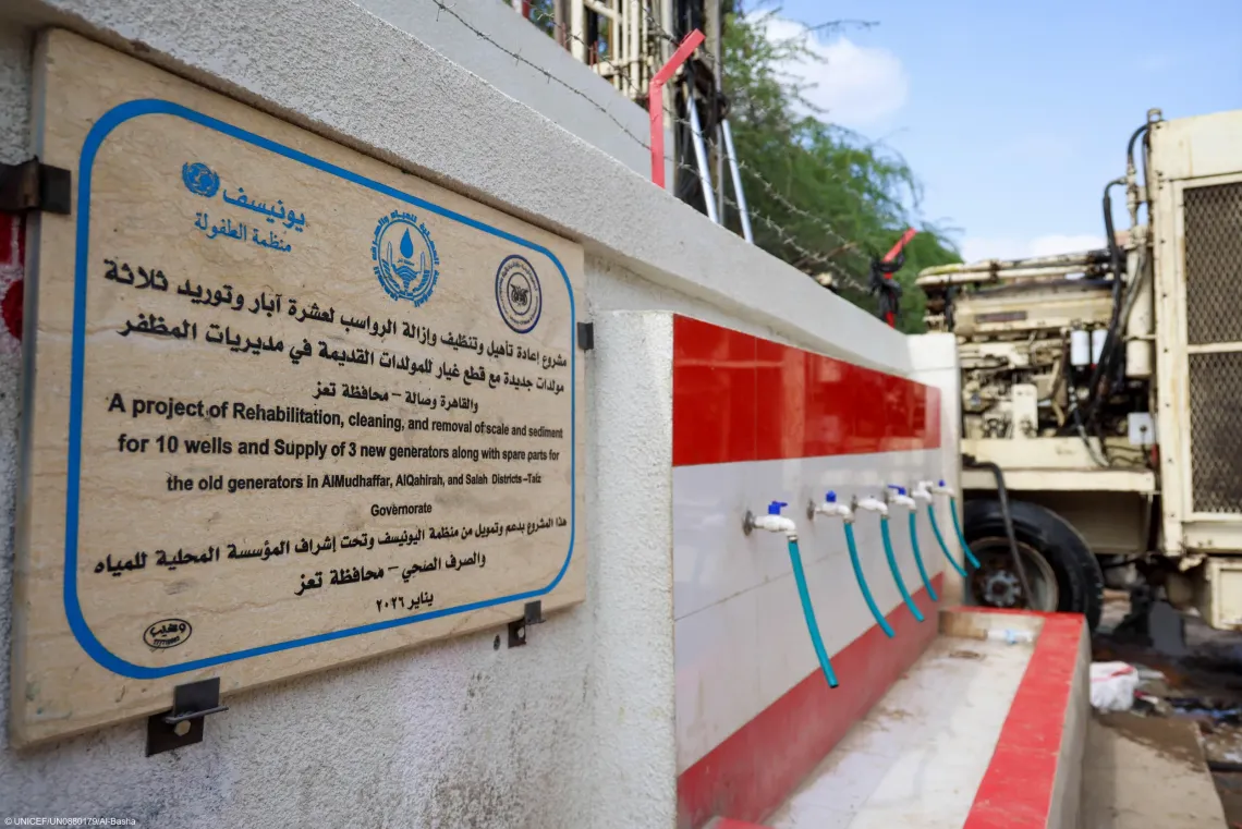 A stone project plaque on a wall next to a multi-faucet public water station and heavy machinery, detailing a UNICEF-supported well rehabilitation project in Taiz Governorate dated January 2026