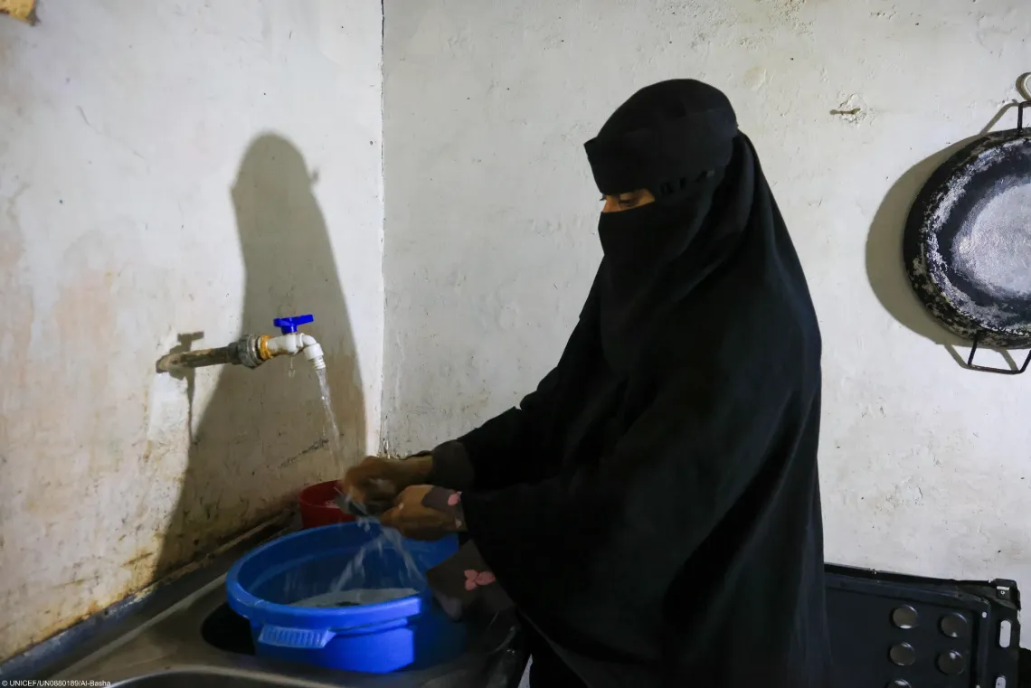 A woman in a black niqab washing items under running tap water into a blue basin set inside a kitchen sink