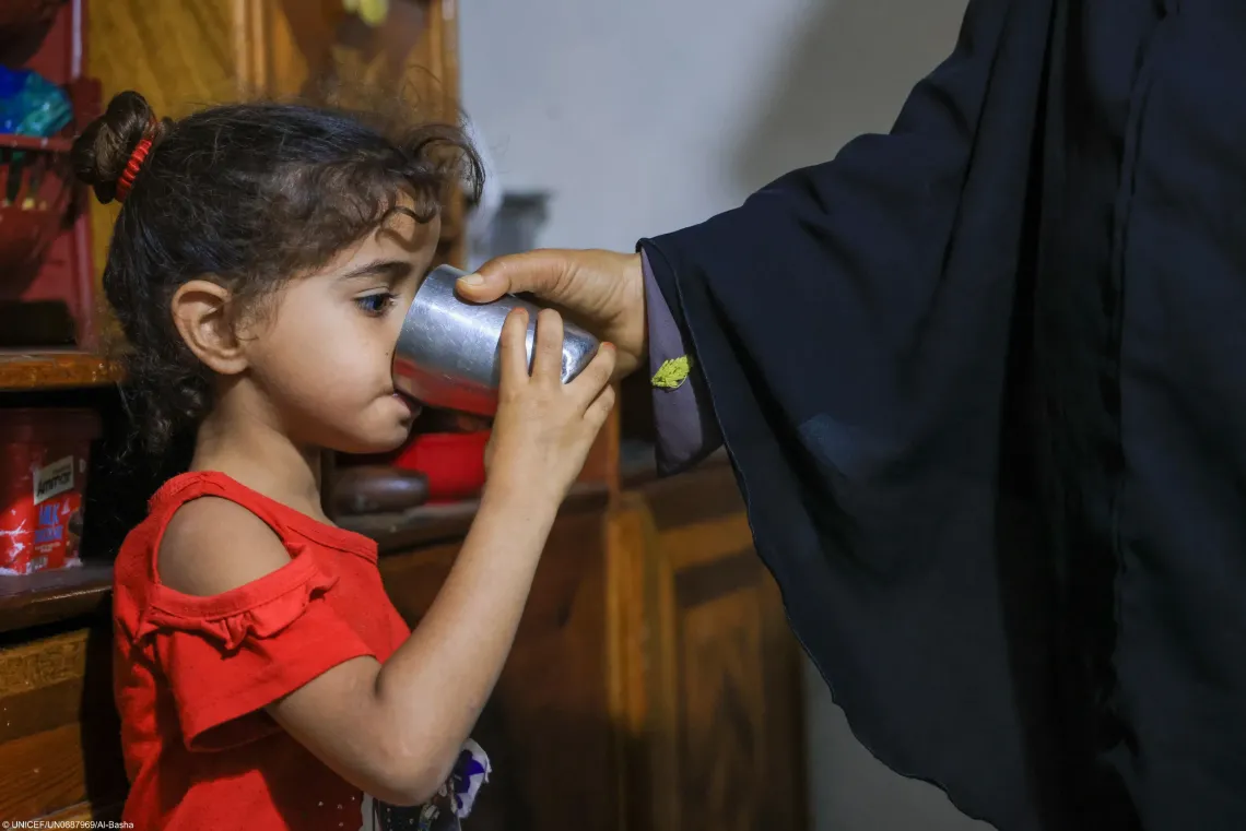 A young girl in a red top drinking water from a metal cup held with the assistance of an adult wearing a black sleeve