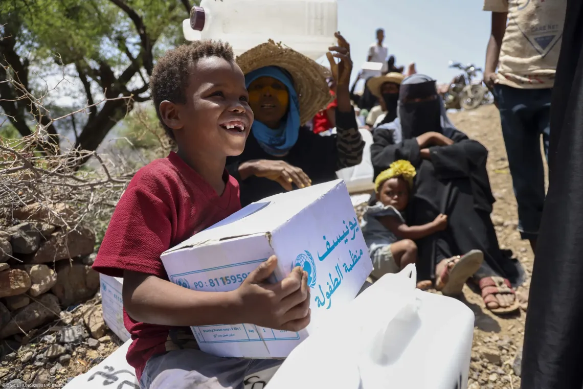 In an outdoor displaced persons site in Yemen, a young boy smiles as he receives a UNICEF relief supply box during a humanitarian aid distribution to support vulnerable families.