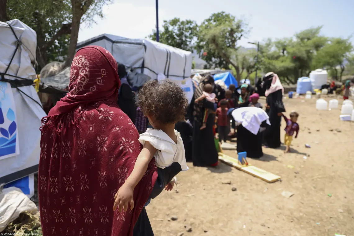 A mother carrying her young child walks through a displaced persons camp in Yemen to access essential humanitarian relief services provided for vulnerable families.