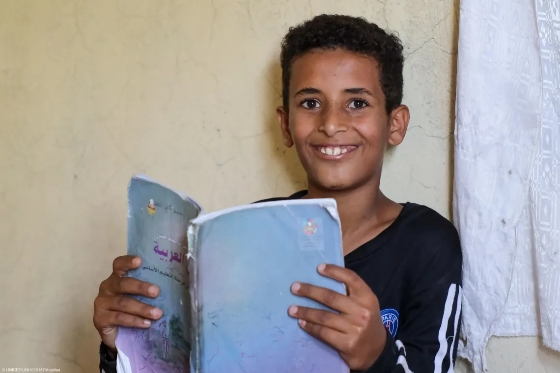 The boy sits indoors holding an open Arabic-language textbook, smiling directly at the camera.
