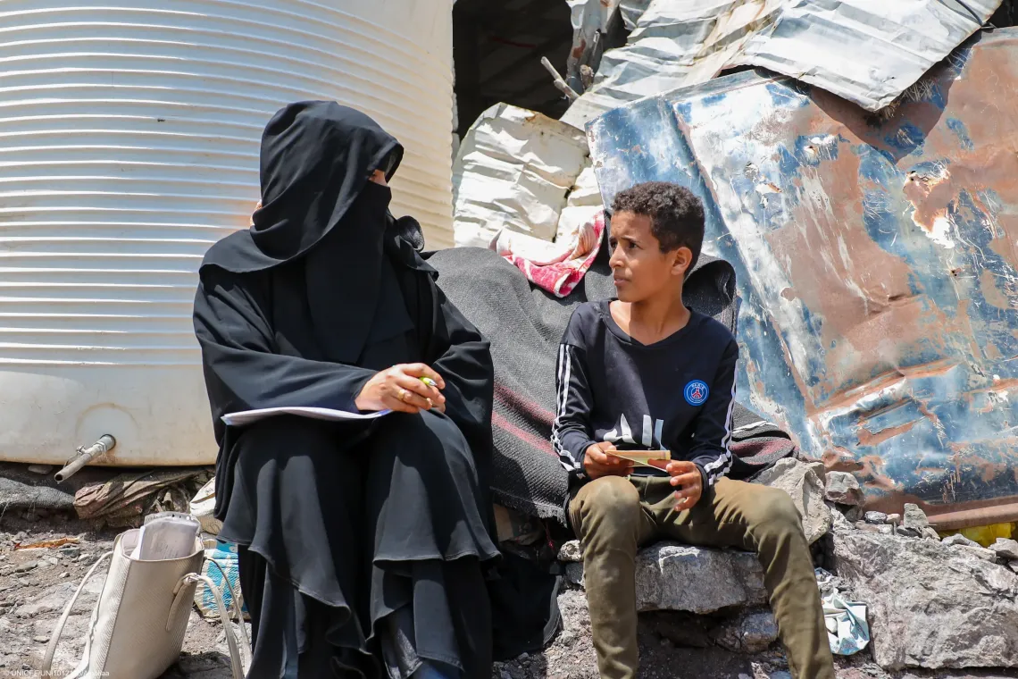 The boy sits on concrete debris next to a woman dressed in a full black niqab and abaya who holds papers and a pen, appearing to conduct an interview or having a conversation.