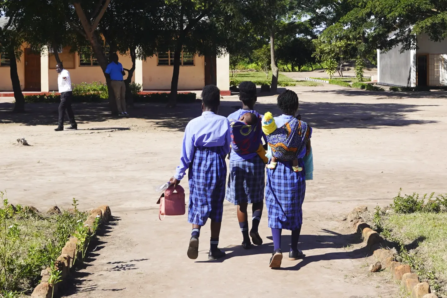 Three learners walk through a school grounds, two carrying young children, at a school in Chifunabuli District, Luapula Province.