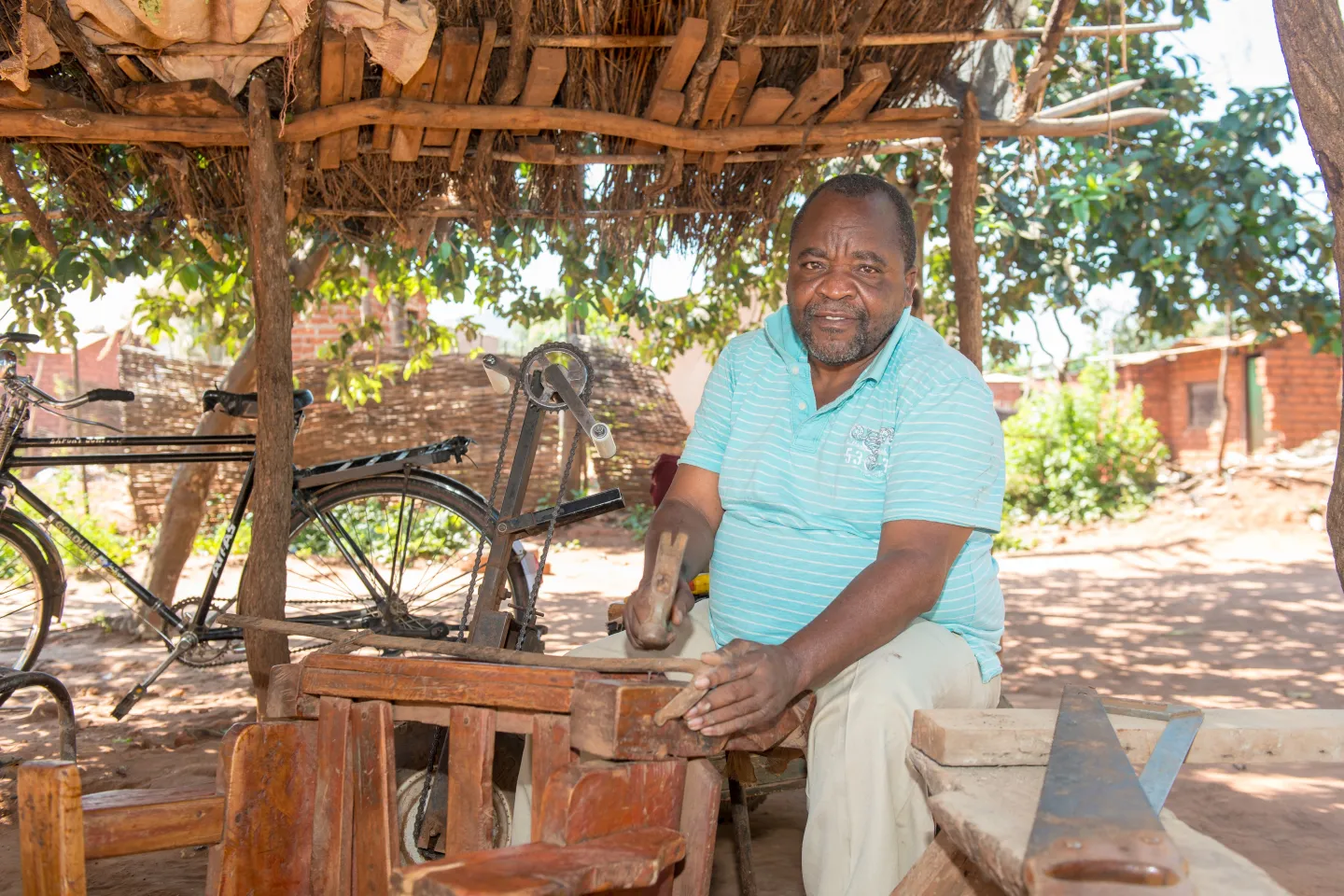 An elderly man sits at a carpentry bench with a bike behind in Zambia