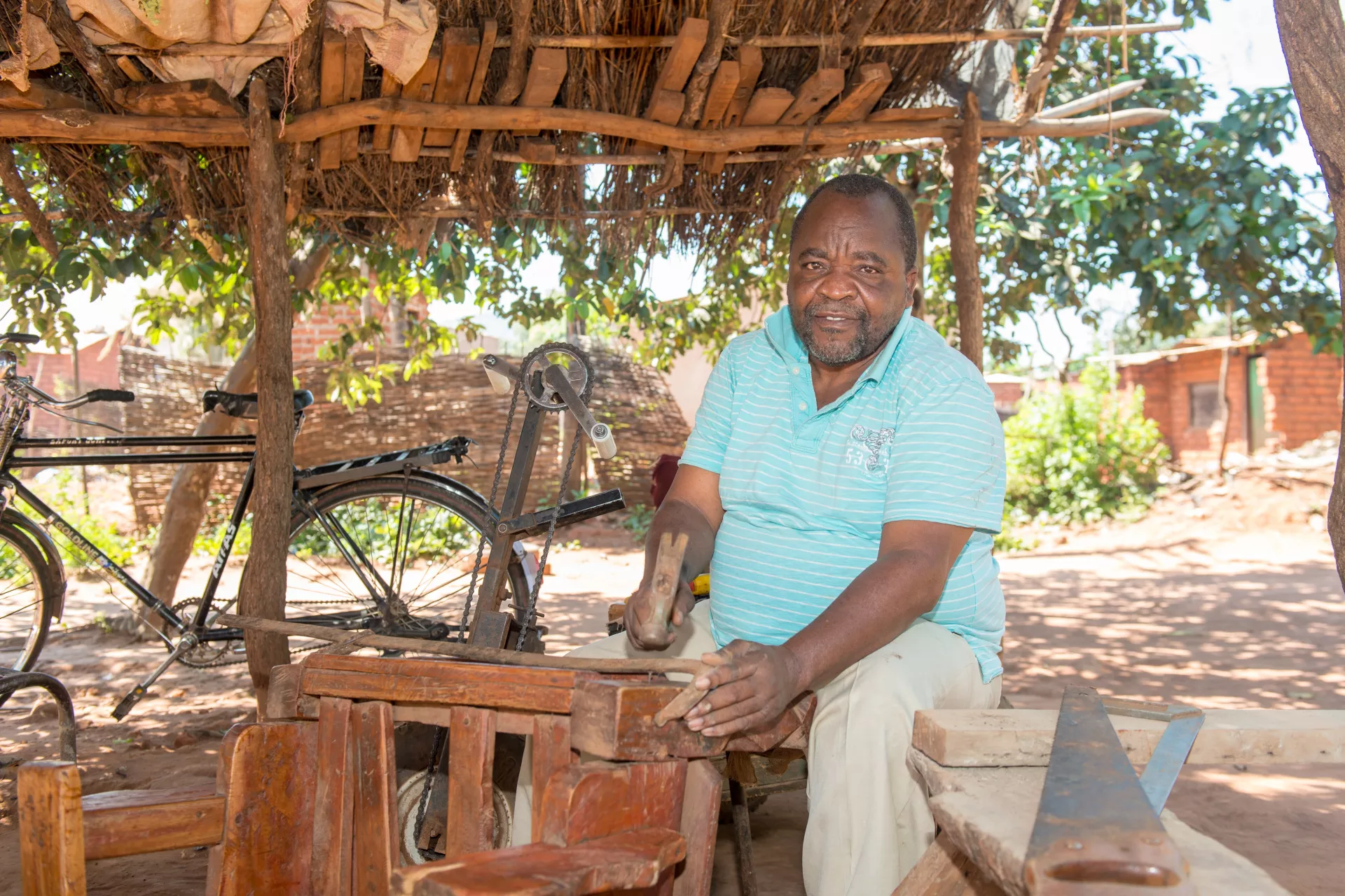 An elderly man sits at a carpentry bench with a bike behind in Zambia