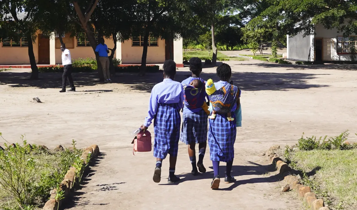 Three learners walk through a school grounds, two carrying young children, at a school in Chifunabuli District, Luapula Province.