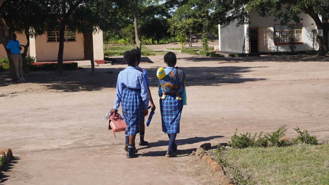 Learners walk across the school grounds, with one carrying a young child.