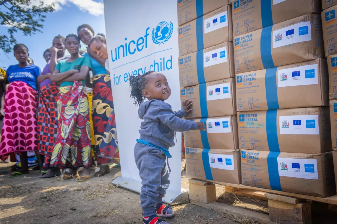 A young child stands next to stacked boxes of Super Cereal Plus, distributed by UNICEF and partners, while a group of women look on in the background. A UNICEF banner with the slogan "for every child" is displayed beside the boxes.