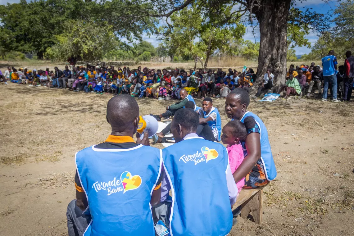 A community meeting takes place outdoors under trees, with a large group of women and children seated in the background. In the foreground, facilitators wearing blue &ldquo;Tikonde Bana&rdquo; vests address the group using a megaphone.