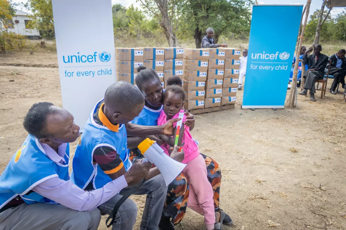 A young girl is screened for malnutrition using a MUAC (mid-upper arm circumference) tape by a group of health workers wearing &ldquo;Tikonde Bana&rdquo; vests. UNICEF banners and stacks of nutrition supply boxes are visible in the background.