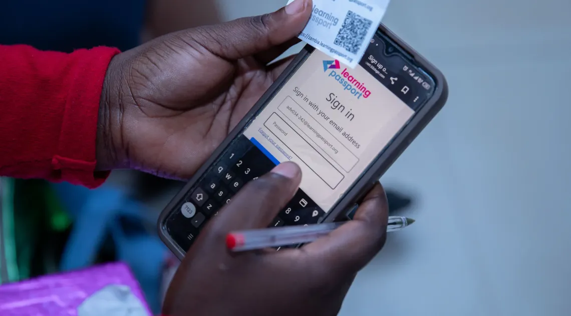 A student logs in to the Learning Passport platform using a smartphone while holding a card with login details and a red pen.